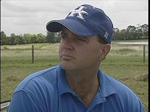 Head shot of John Hancock wearing UK baseball cap with farm field in background.