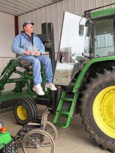Keith Posselt sits, approximately six feet in the air, on seat of lift attached to tractor that raises him to the cab. His wheelchair sits on floor in foreground.