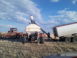 Man standing on grain cart with another man on ground next to semi with tractor in background