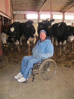 Keith Posselt, dressed in jeans and blue jacket, sits in wheelchair in cattle barn. Five Holstein cows stand behind him behind a wire fence looking at the camera.