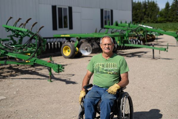 Doug sits in wheelchair with farm equipment and barn behind him