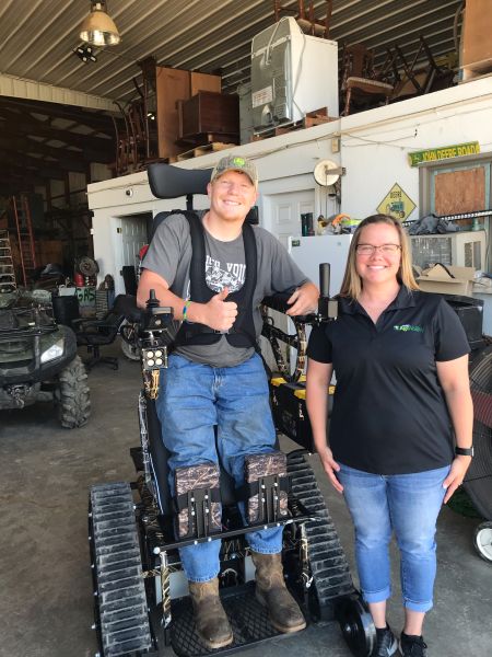 Kane in stand-up tracked wheelchair giving thumbs up and standing next to Rachel Jarman in farm shop.