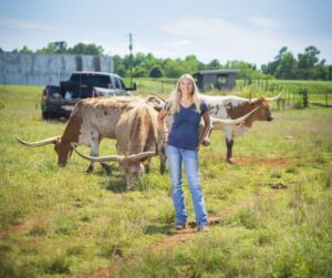 Kristi stands in pasture with three longhorn cattle and a pickup truck behind her.