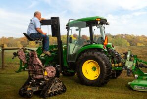 Todd using lift to access cab of tractor with tracked wheelchair on ground near him.