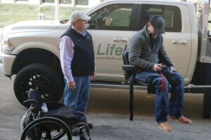 Man sits in lift to raise him into equipment while other man looks on. Wheelchair in foreground.