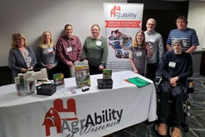 Four women and four men stand behind conference display table looking at camera. One man is in wheelchair.