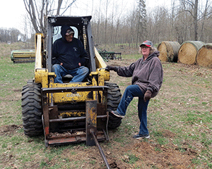 Doug Jones sits in seat of skid-steer loader while Sonja Jones stands with foot on loader and hand on grab bar on loader